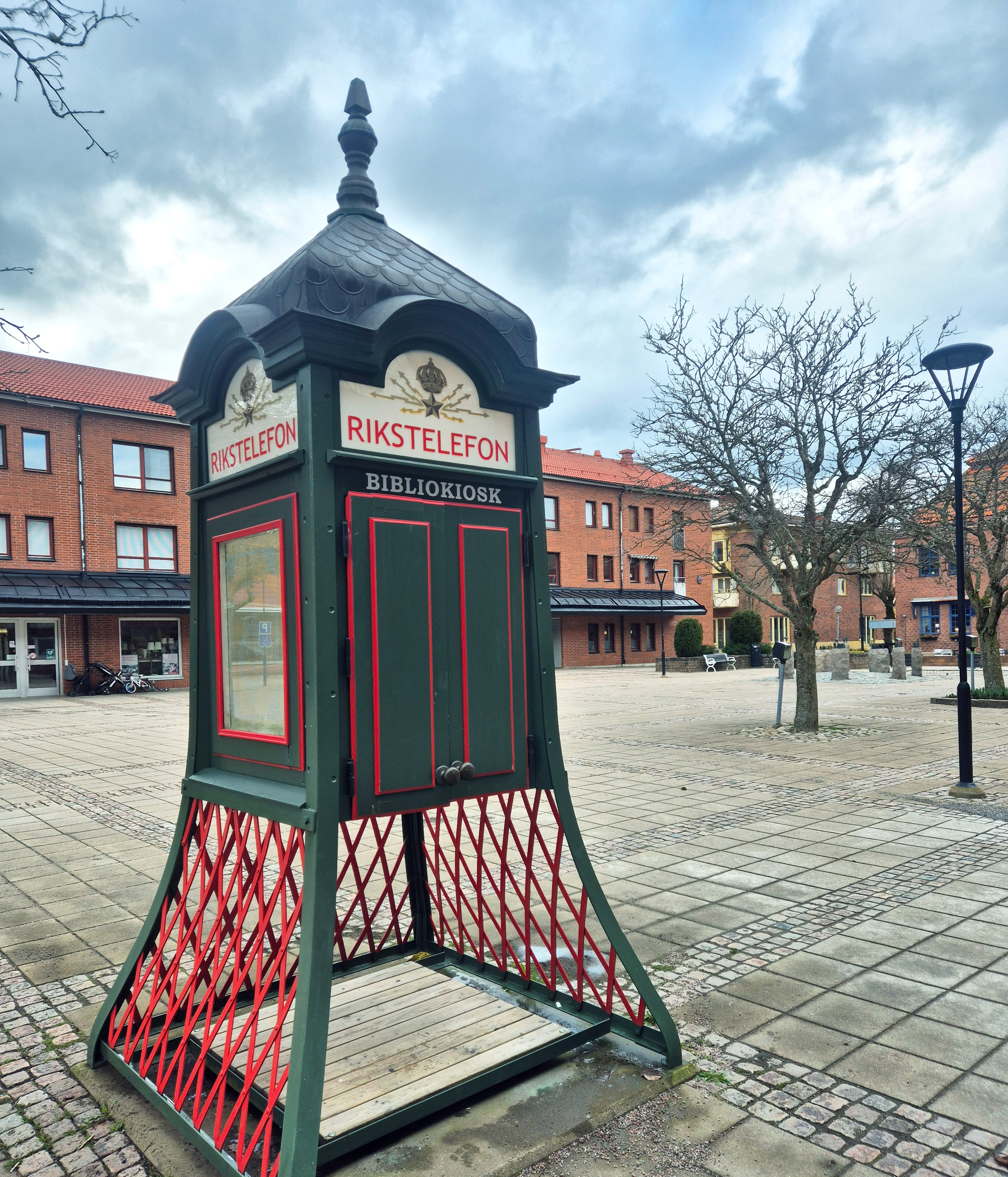 Bilden visar rikstelefonkiosken på torget i Skillingaryd, en grön telefonkiosk med röda detaljer. Ovanför dörrarna står det "Bibliokiosk" med vita bokstäver.